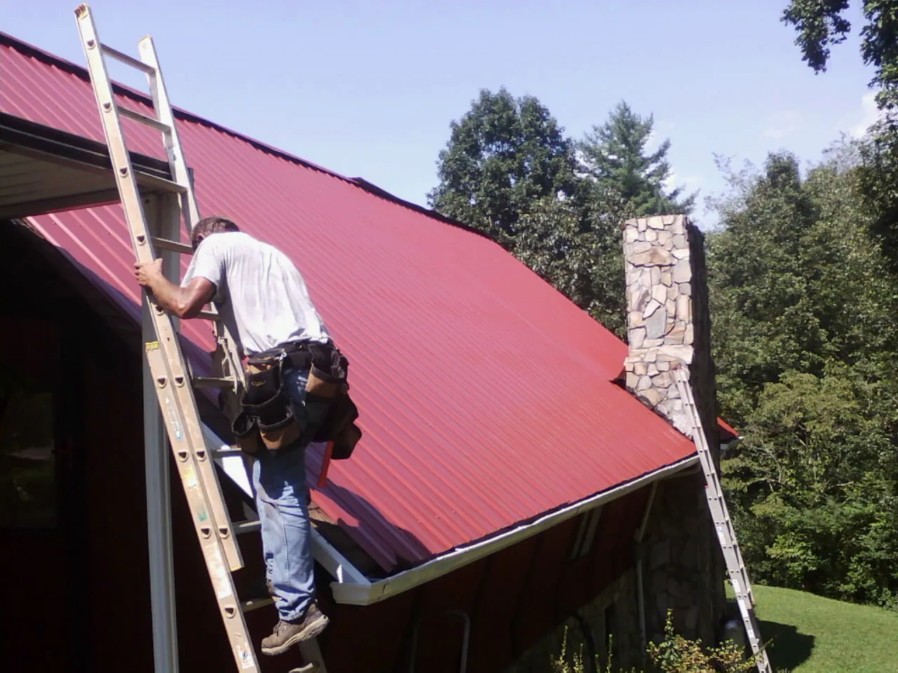 Roofer on a ladder beside a red metal roof and stone chimney — Soffit & Fascia Repair by Roofing Brothers in Ardara